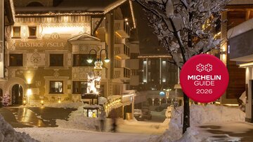 Ein malerisches Winterdorf mit verschneiten Gebäuden und festlicher Beleuchtung. Im Vordergrund ist das Logo des Michelin Guides 2026 zu sehen.